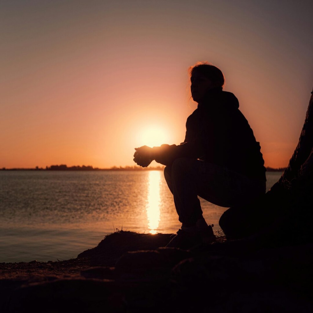 Person sitting down on a rock overlooking the ocean at sunset, representing the psychological effects of environmental loss. 